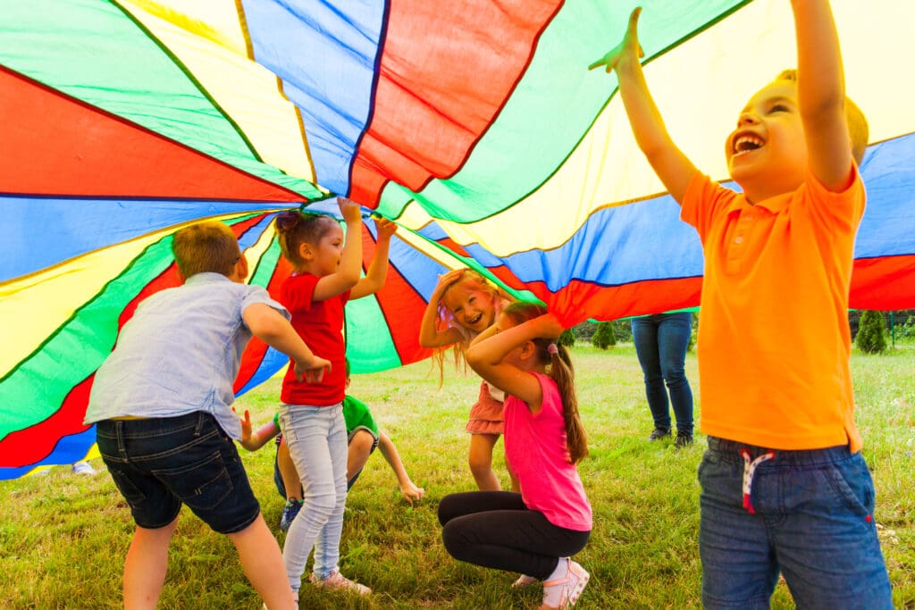 Children laugh and play under a colorful parachute during an outdoor activity, enjoying teamwork and fun in the sun.